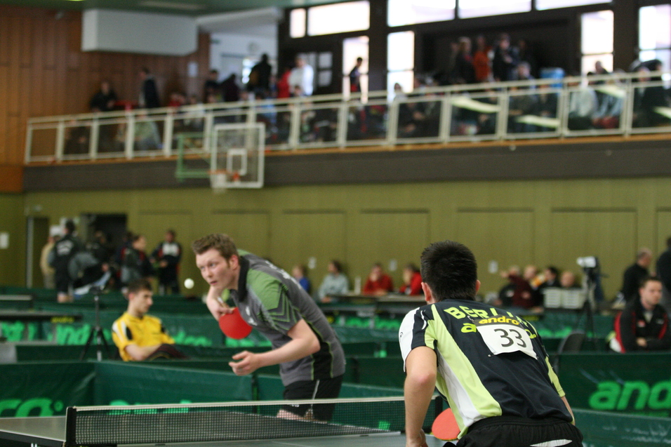 Zwei Tischtennisspieler stehen sich gegenüber, einer beim Aufschlag, umgeben von Zuschauern in einer Sporthalle.