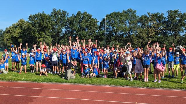 Gruppe von Kindern in blauen T-Shirts mit roten Mützen hebt die Hände auf einem Sportplatz unter blauem Himmel.