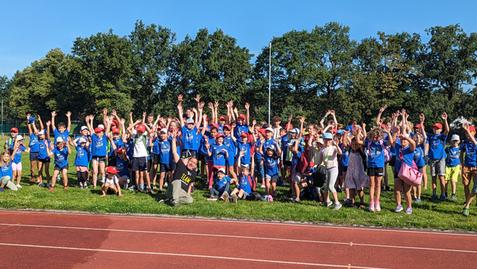 Gruppe von Kindern in blauen T-Shirts mit roten Mützen hebt die Hände auf einem Sportplatz unter blauem Himmel.