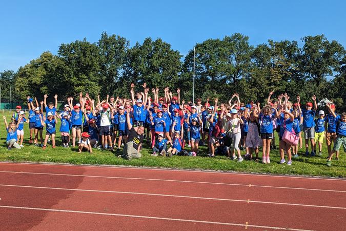 Gruppe von Kindern in blauen T-Shirts mit roten M&uuml;tzen hebt die H&auml;nde auf einem Sportplatz unter blauem Himmel.