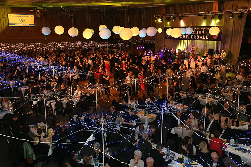 Blick auf eine festliche Ballveranstaltung mit Gästen, beleuchteten Tischen und einer Band auf der Bühne.