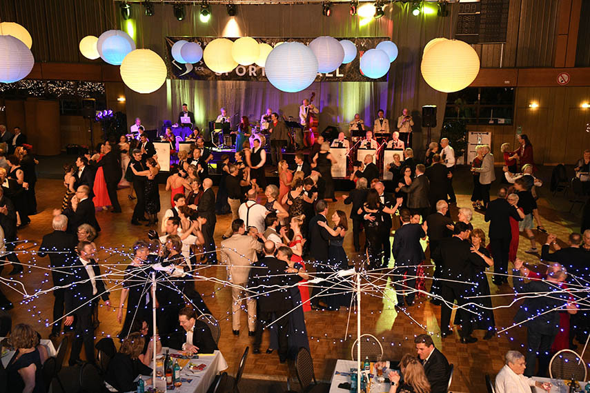 Tanzende Menschen auf einem Ball, umgeben von dekorativen Laternen und einer Live-Band im Hintergrund.