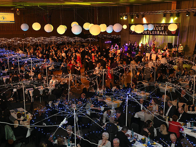 Blick auf eine festliche Ballveranstaltung mit Gästen, beleuchteten Tischen und einer Band auf der Bühne.