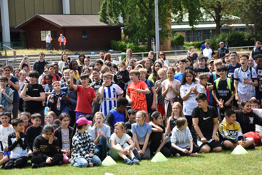 Gruppe von Kindern und Jugendlichen versammelt auf einem Sportplatz, einige winken und lächeln in die Kamera.
