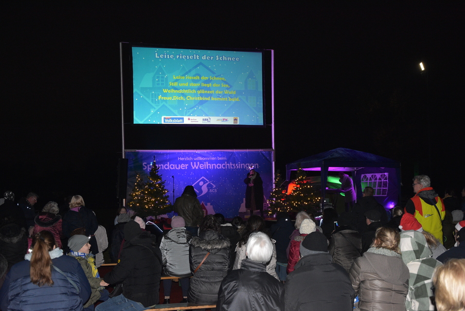 Menschen versammeln sich bei einer Weihnachtsveranstaltung, ein Bildschirm zeigt den Text „Leise rieselt der Schnee“.