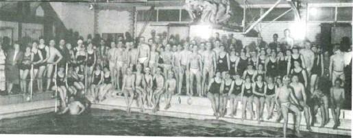 Schwarz-weiß Aufnahme einer großen Gruppe von Schwimmern in Badesachen am Rand eines Hallenbades.