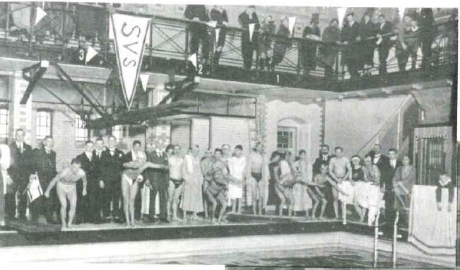 Schwarz-weiß Foto einer Schwimmveranstaltung mit Athleten am Beckenrand und Zuschauern auf der Galerie.