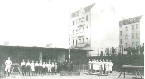 Gruppenaufnahme von Sportlern in historischen Uniformen vor einem Übungsplatz mit Geräten und Wohngebäuden im Hintergrund.