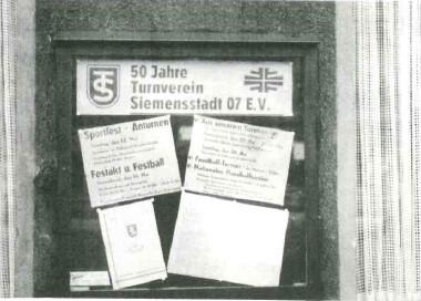 Schild und Poster in einem Fenster, die das 50-jährige Bestehen des Turnvereins Siemensstadt 07 E.V. ankündigen.