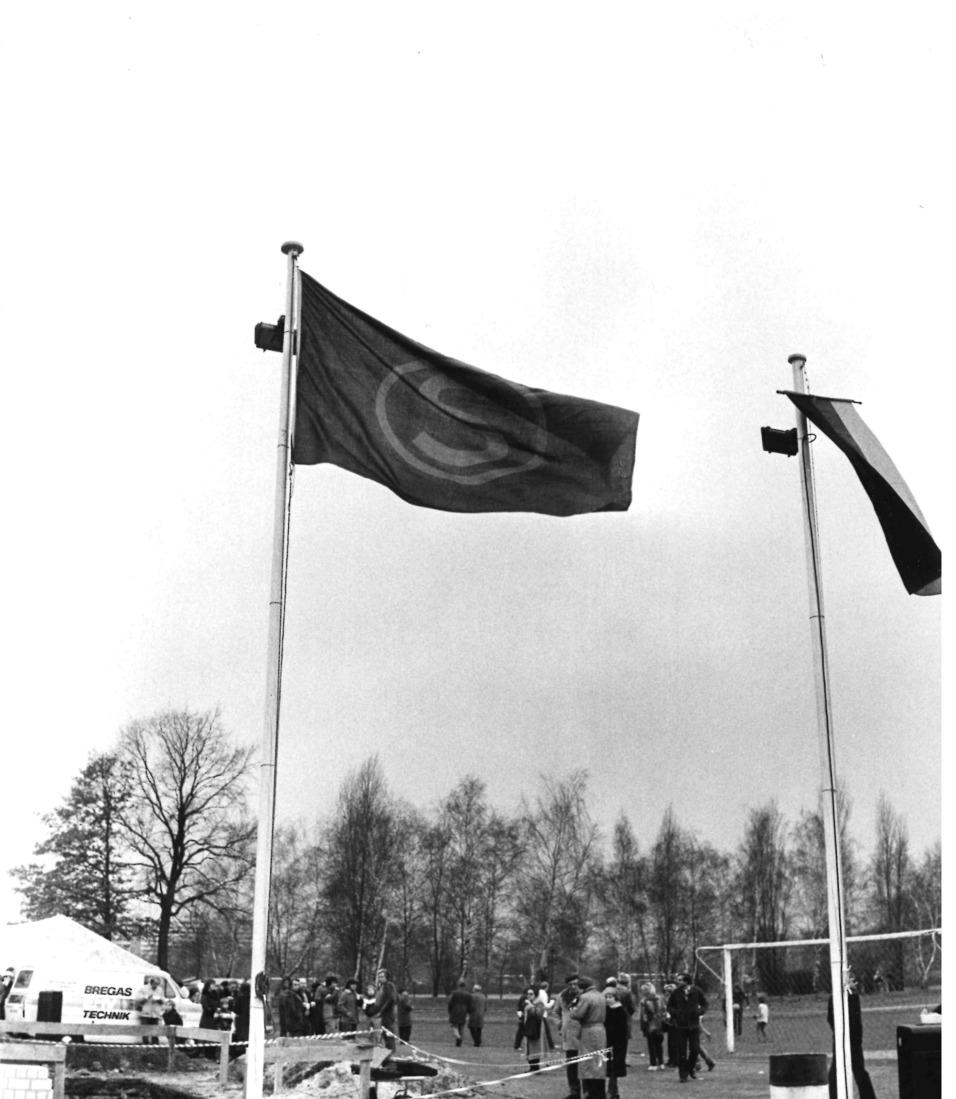 Zwei Fahnen wehen an Masten vor einer Menschenmenge und einem Zelt in einer Sportumgebung.