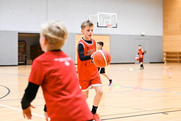 Junge in rotem Basketballtrikot dribbelt einen Ball in einer Sporthalle, während andere Kinder trainieren.