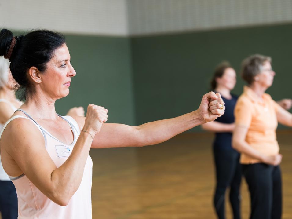 Frau in Sportkleidung führt Bewegungen in einer Gruppe bei einem Fitnesskurs durch, fokussiert auf die Armhaltung.