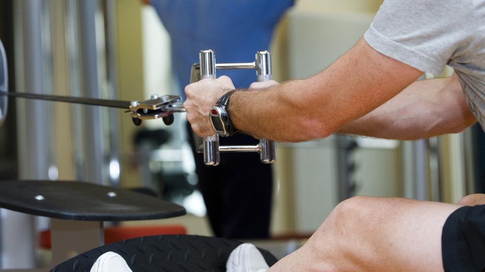 Mann trainiert im Fitnessstudio mit einem Kabelzuggerät, ein anderer Mensch im Hintergrund führt eine Übung aus.