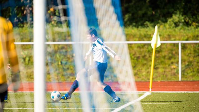 Wettkampf auf dem Fußballplatz: Ein Spieler in weiß-blauer Trikotausstattung schießt einen Ball ins Spiel.