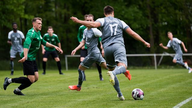 Drei Fußballspieler in grünen und grauen Trikots auf einem Rasenplatz, während ein Spieler den Ball zuspielt.