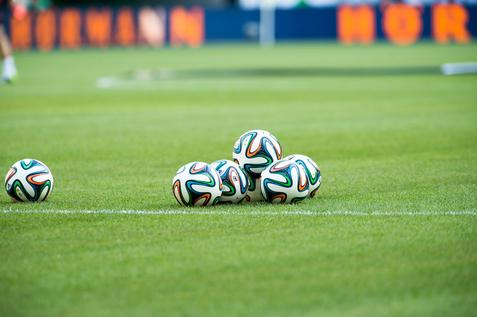 Fünf Fußballbälle liegen auf einem grünen Rasenfeld, sichtbar sind auch der Spielfeldrand und eine verschwommene Hintergrundszene.
