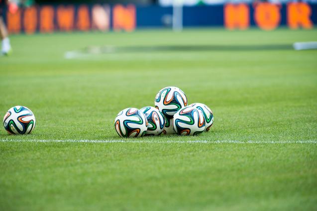 Fünf Fußballbälle liegen auf einem grünen Rasenfeld, sichtbar sind auch der Spielfeldrand und eine verschwommene Hintergrundszene.