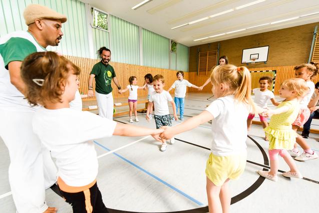 Gruppe von Kindern in T-Shirts und Erwachsenen in einer Turnhalle, die gemeinsam einen Kreis bilden und spielen.
