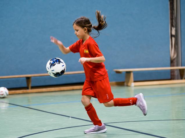 Mädchen in rotem Trikot dribbelt einen Fußball in einer Halle mit grünem Boden und blauen Wänden.