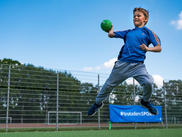 Junge springt auf einem Sportplatz und wirft einen grünen Handball, umgeben von einem Zaun und Bäumen im Hintergrund.