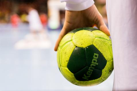 Nahaufnahme einer Hand, die einen grünen Handball mit gelben Elementen hält, im Hintergrund Spieler auf dem Spielfeld.