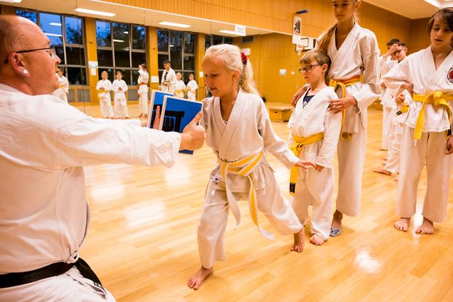 Trainer überreicht einem Mädchen in Karate-Gi mit gelbem Gürtel zwei blaue Urkunden während des Trainings.