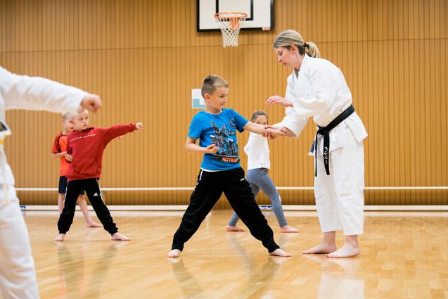 Kinder üben Karate in einem Trainingsraum mit einem Lehrer in weißem Gi und schwarzem Gürtel.