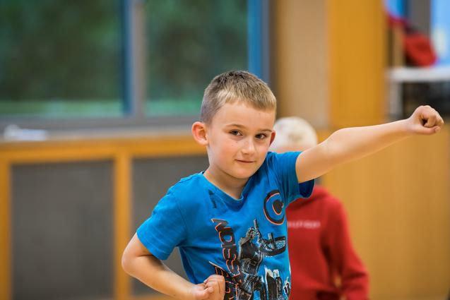Junge im blauen T-Shirt zeigt mit erhobener Hand einen kraftvollen Ausdruck in einem hellen Raum.