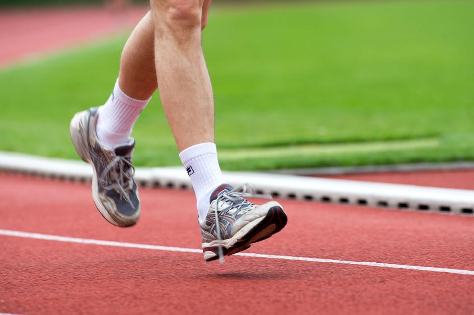Läufer in grauen Sportschuhen hebt den Fuß auf einer roten Laufbahn über grünem Rasen.
