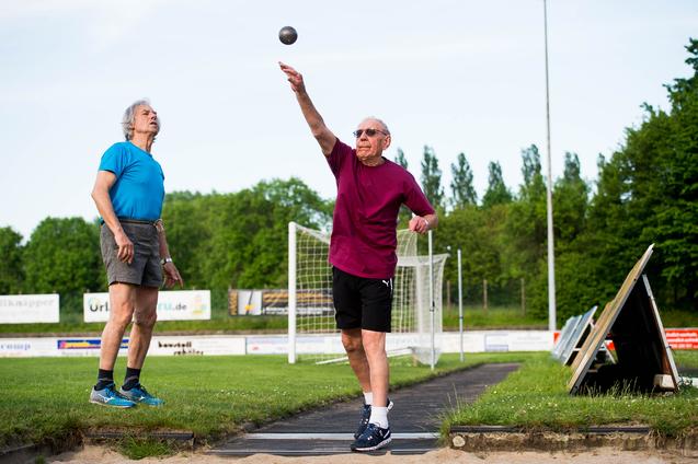 Zwei ältere Männer üben das Kugelstoßen auf einem Sportplatz, einer wirft die Kugel, der andere beobachtet.