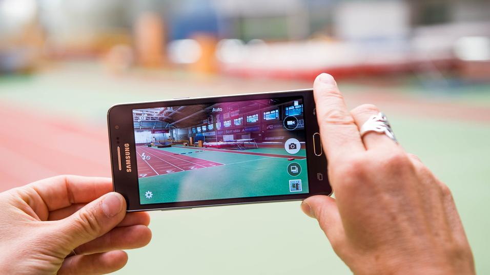 Hand hält ein Smartphone, das eine Sporthalle mit einer Laufbahn im Hintergrund zeigt.