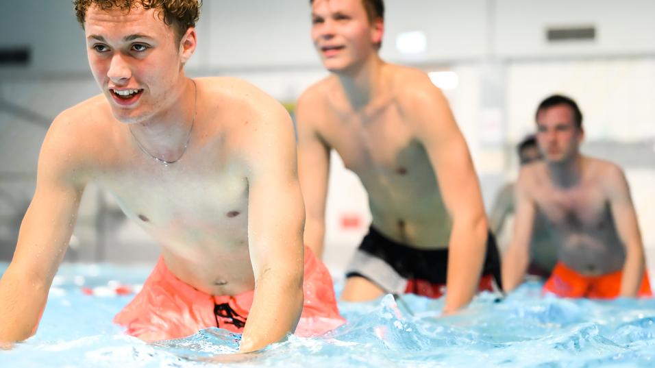 Vier junge Männer in Badebekleidung üben im Wasser eines Schwimmbeckens, einige lachen und spritzen.