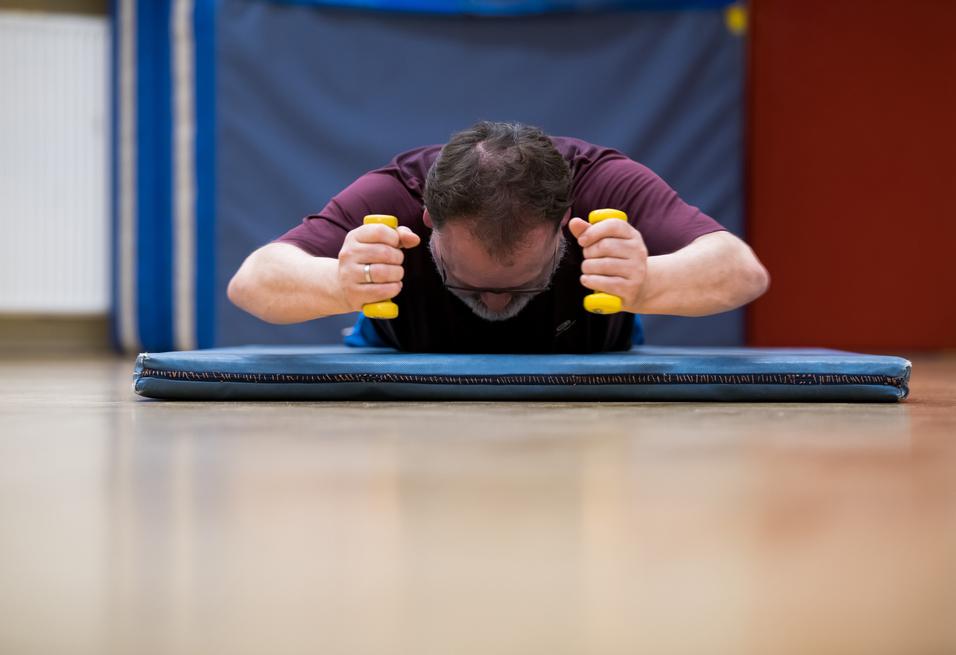 Mann trainiert auf einer Matte mit gelben Hanteln, Kopf nach unten, in einem Sportraum mit blauen Wänden.