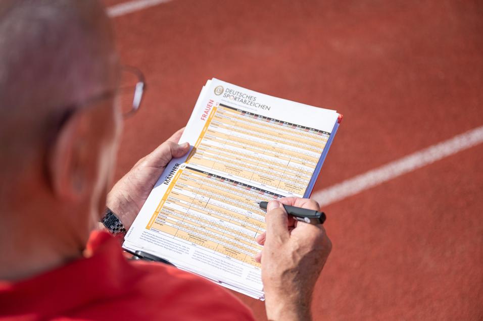 Hand eines Prüfers, der auf einem Blatt mit Sportabzeichen-Anforderungen auf einer Laufbahn notiert.