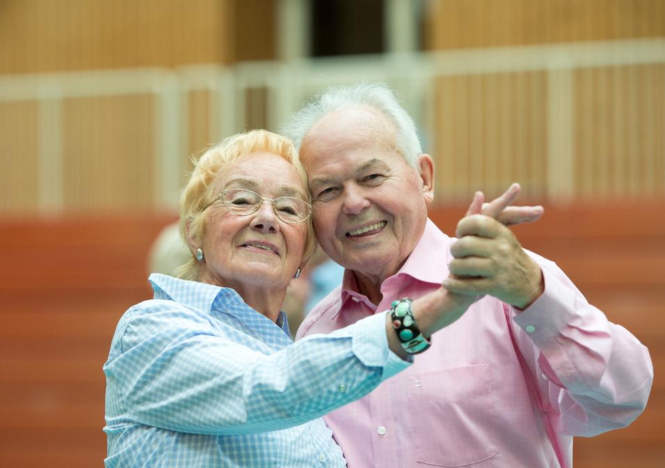 Ein älteres Paar umarmt sich lächelnd und tanzt in einer Sporthalle. Ihre Kleidung ist hell und farbenfroh.
