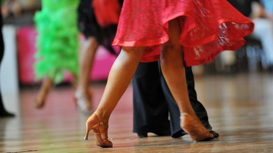 Tanzende Beine in eleganten Schuhen, während sich ein rotes Kleid dreht, vor unscharfem Hintergrund.