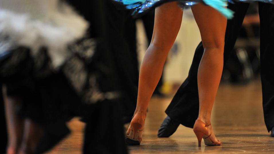 Tanzende Beine in eleganten Schuhen, umgeben von schwingenden Kostümen auf einem Holzboden.