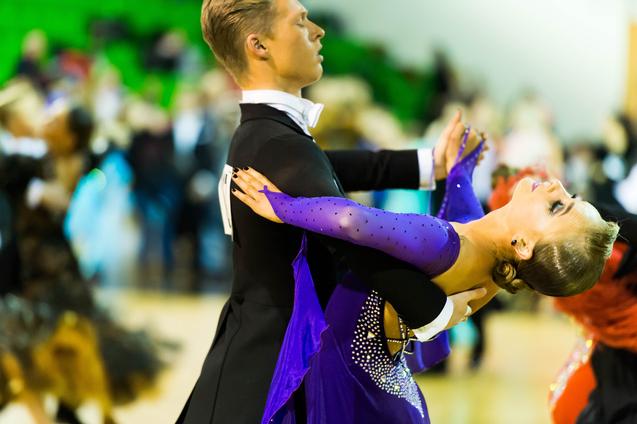 Tanzpaar in eleganter Haltung auf der Tanzfläche, die Frau trägt ein glitzerndes lila Kleid und lächelt.