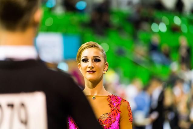 Tänzerin mit blondem Haar und glitzerndem pinken Kleid blickt konzentriert zu ihrem Partner bei einem Wettkampf.