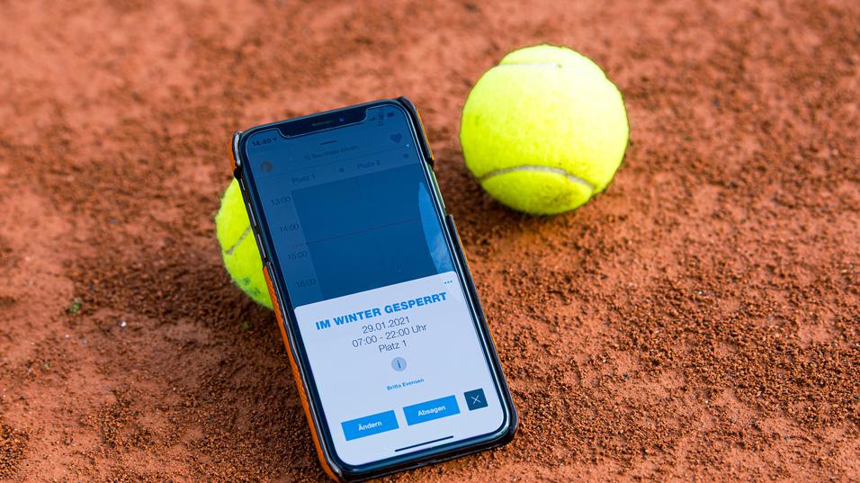 Smartphone mit Buchungsinfo liegt auf einem Tennisplatz, neben zwei gelben Tennisbällen auf rotem Sand.