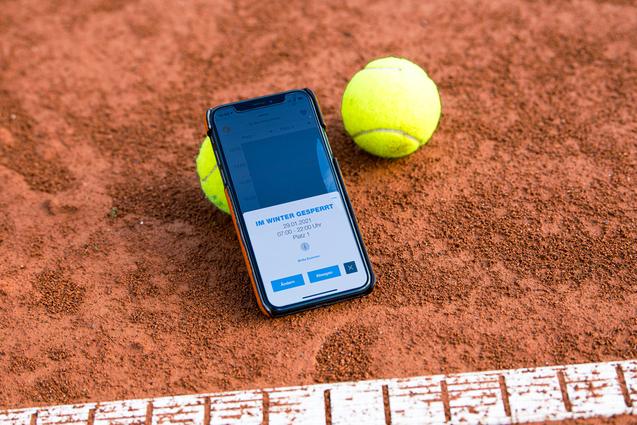 Smartphone mit Buchungsinfo liegt auf einem Tennisplatz, neben zwei gelben Tennisbällen auf rotem Sand.