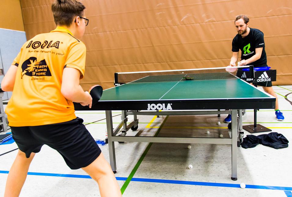 Zwei Tischtennisspieler trainieren an einem Tischtennistisch in einer Sporthalle, mehrere Bälle liegen auf dem Boden.