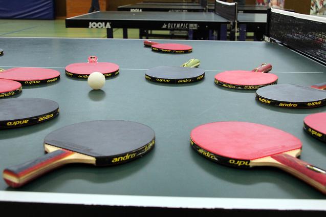 Mehrere Tischtennisschläger in verschiedenen Farben und ein Tischtennisball liegen auf einer grünen Tischtennisplatte.