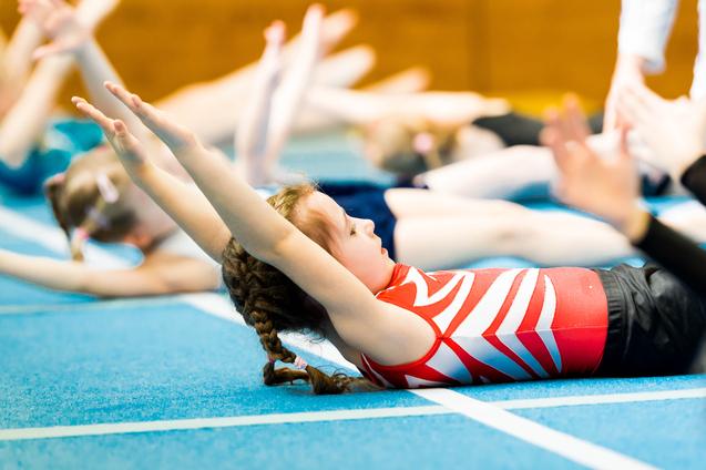 Mädchen mit Zöpfen liegt auf einer blauen Gymnastikmatte und hebt die Arme während eines Trainings.