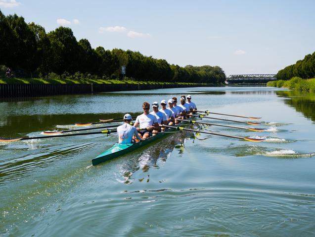 Acht Ruderer in weißen Shirts rudern synchron auf einem ruhigen Gewässer, umgeben von Bäumen und einem blauen Himmel.