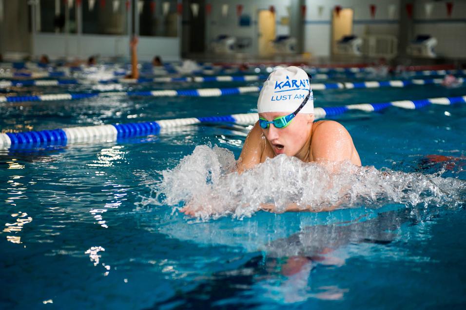 Schwimmerin in Aktion beim Brustschwimmen in einem Innenbecken, mit spritzendem Wasser und Schwimmbrille.