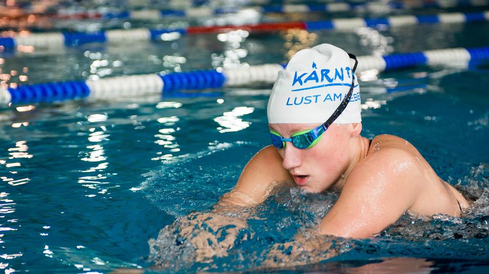 Schwimmerin in blauer Badekappe und Schwimmbrille, die im Wettkampfbecken krault. Spritzer umgeben sie.