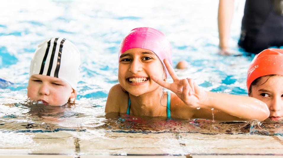 Lächelndes Mädchen mit pinker Badehaube zeigt V-Zeichen im Wasser; zwei weitere Kinder sind im Hintergrund zu sehen.