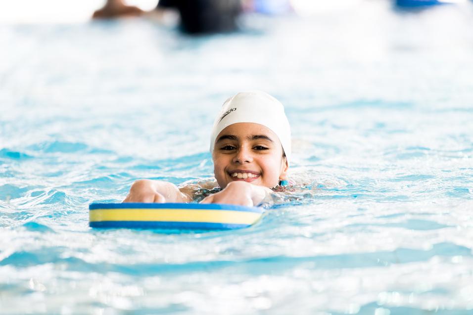 Lächelndes Kind mit Badekappe schwimmt auf einem Schwimmbrett im Wasser eines Hallenbades.