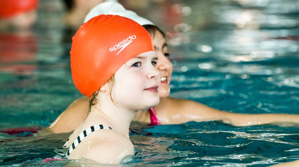Mädchen mit oranger Schwimmhaube schwimmt in einem Hallenbad, im Hintergrund weitere Kinder im Wasser.